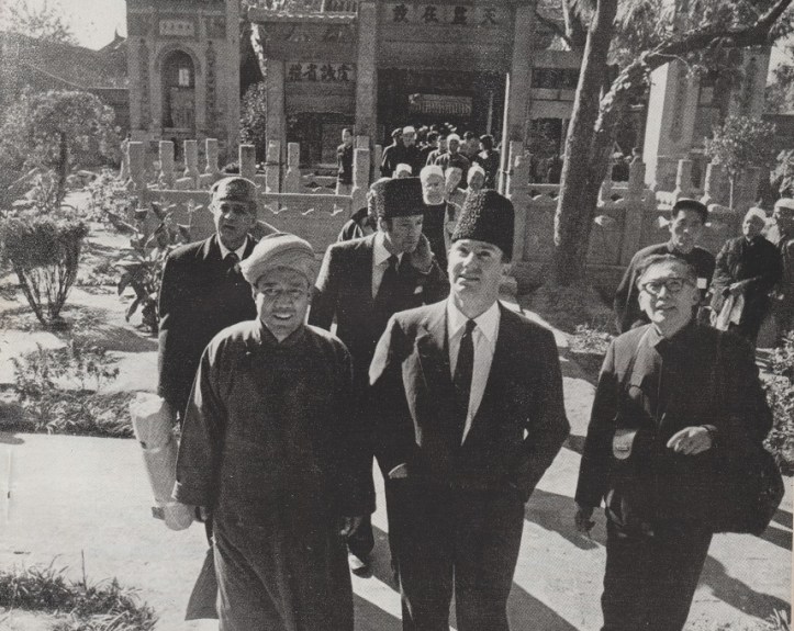 Historical photograph featuring several men walking together in a garden setting, with traditional attire and hats. A crowd is visible in the background, and a building is partly visible behind them.