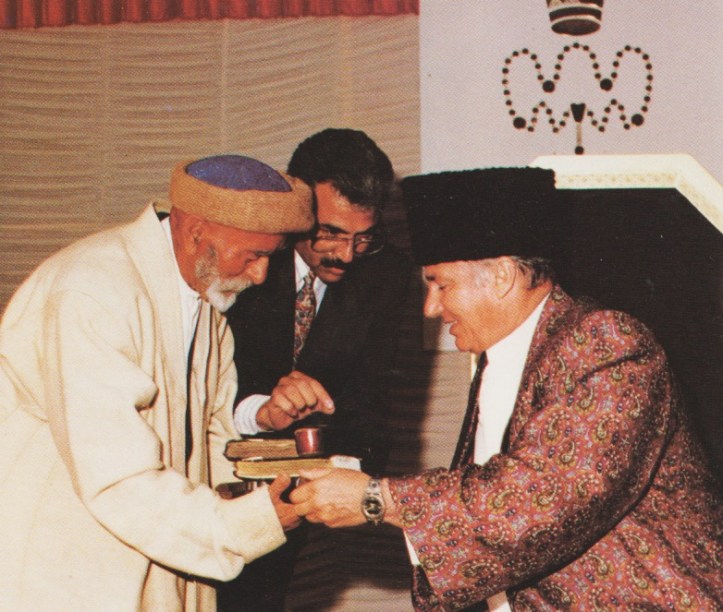 A historic moment featuring three men, one in traditional attire presenting a book to another man in a patterned coat, while a third man observes attentively.