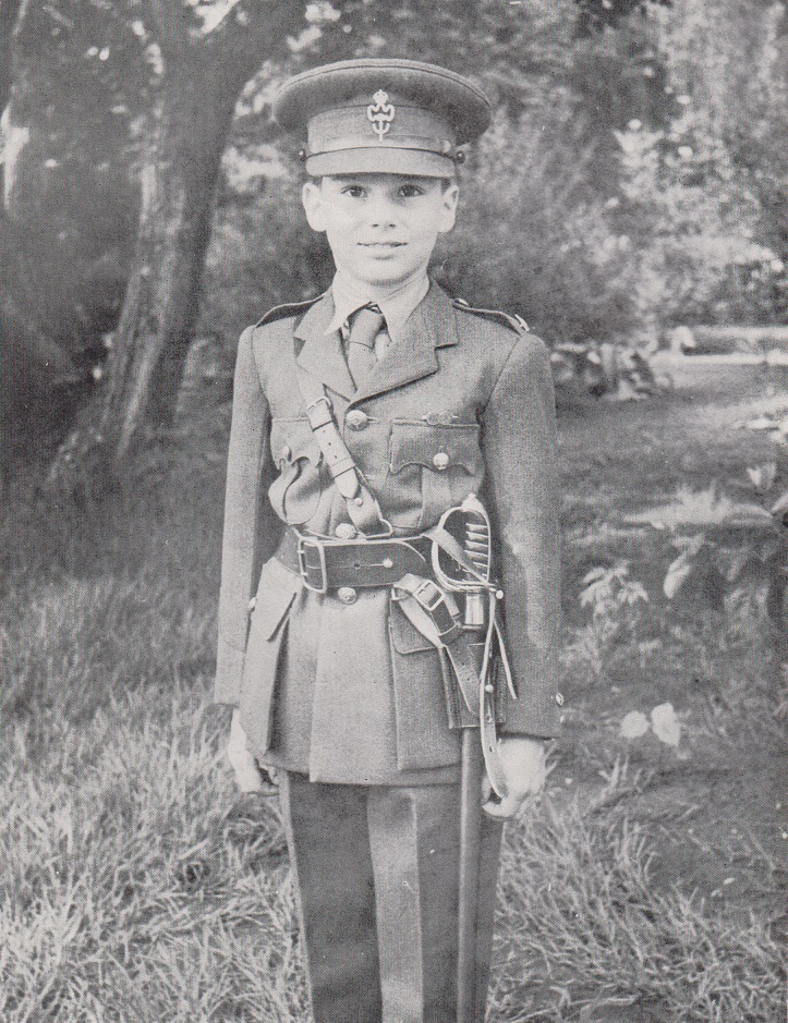 A young boy in a military uniform, complete with a cap and belt, stands confidently in a garden setting, smiling at the camera.