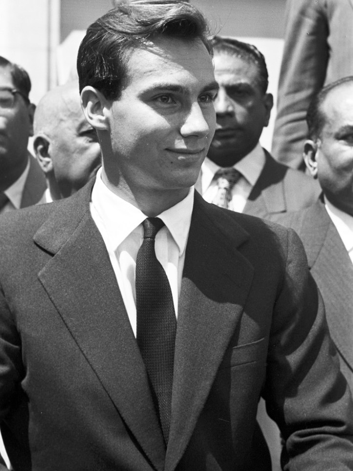 A young man in a black suit and tie smiles while standing in front of a group of men, possibly during a formal event or ceremony.