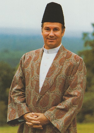 A portrait of Mawlana Shah Karim, His Highness the Aga Khan IV, dressed in traditional attire with a carved hat, standing outdoors with a blurred landscape in the background.