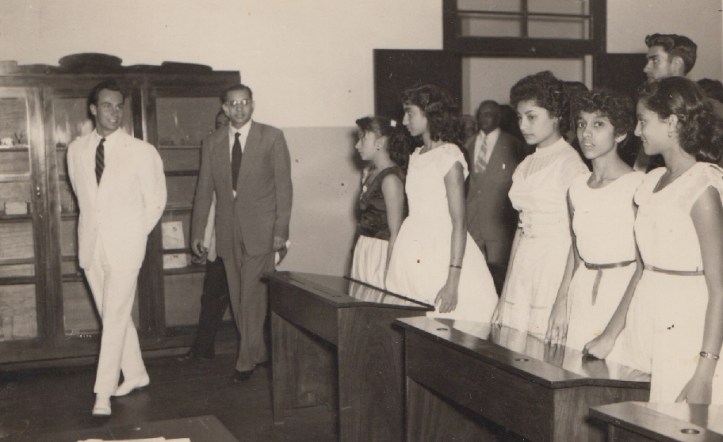 A historic black and white photograph showing a group of young women in white dresses standing at a classroom setting as two men walk past them, one in a white suit.