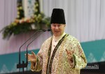 A man in traditional attire leads a speech or presentation at a ceremonial event, with a floral backdrop and microphone setup.