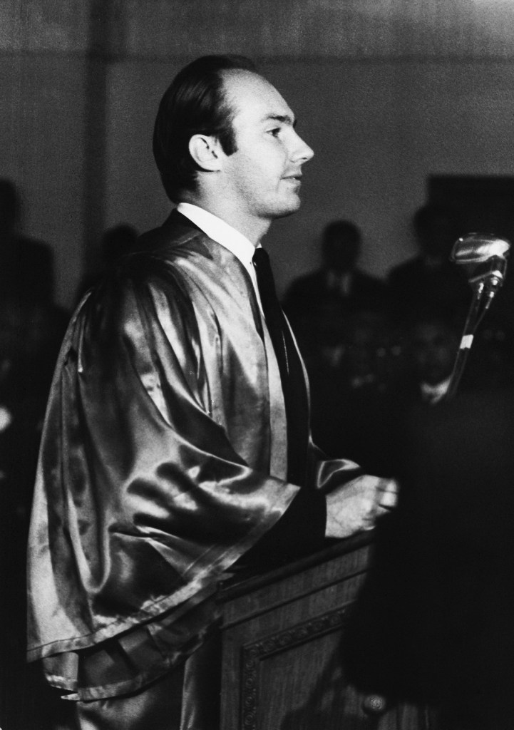 A black and white photograph of a man in academic robes speaking at a podium. The setting appears to be formal with a microphone in front of him and an audience visible in the background.