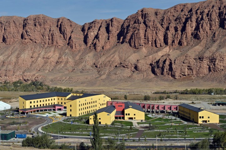 A modern educational facility nestled against a backdrop of rugged red mountains, showcasing colorful architecture with yellow and red buildings surrounded by landscaped gardens.
