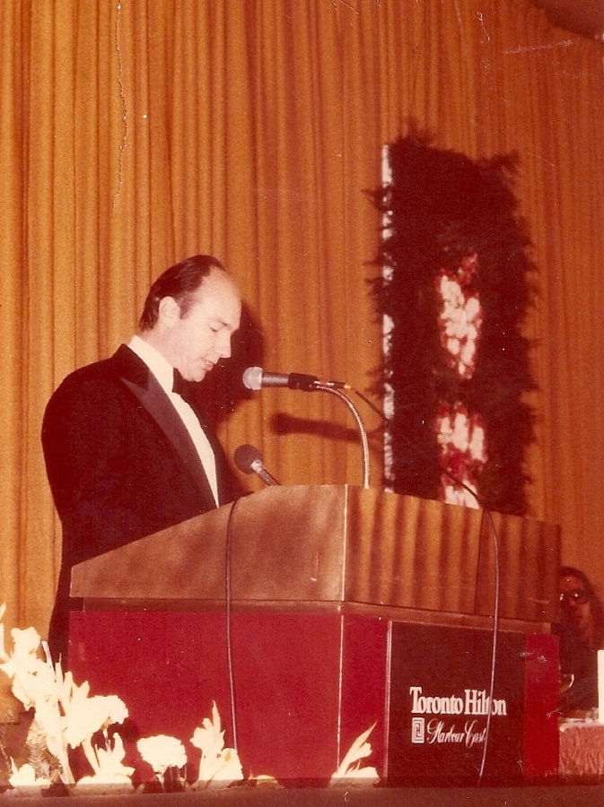 A man in formal attire speaks at a podium decorated with flowers at the Toronto Hilton hotel, with a curtain backdrop.