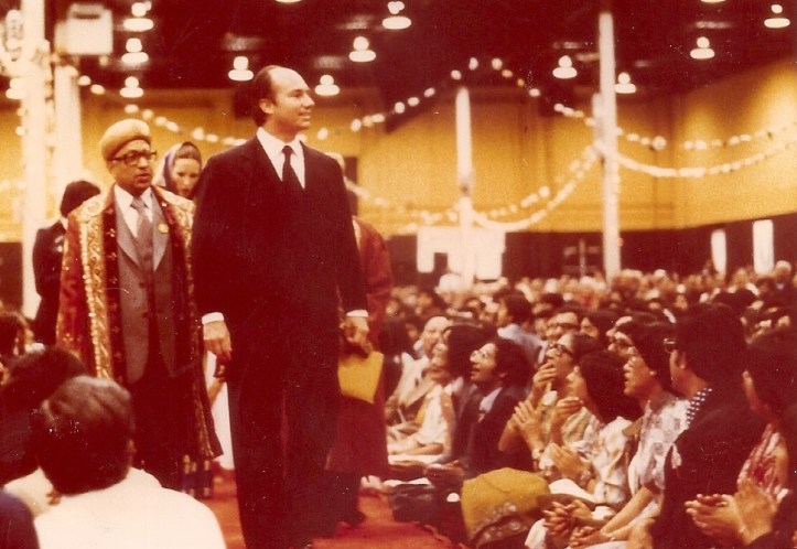 Mawlana Shah Karim walking among an audience during a gathering, dressed in a suit, while a man in traditional clothing stands beside him.