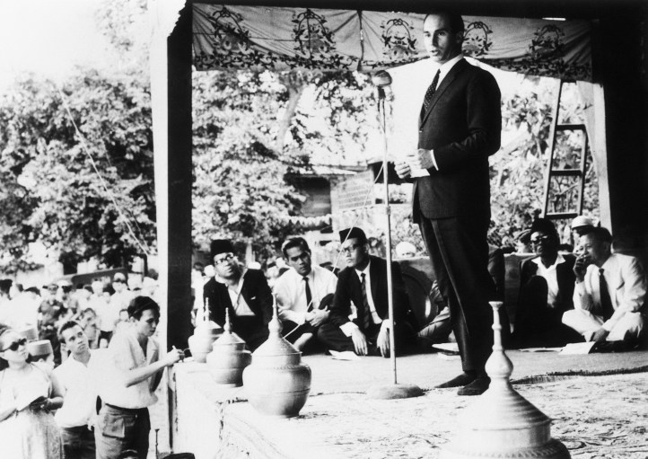 Black and white photograph of Mawlana Shah Karim delivering a speech at an outdoor event, surrounded by an attentive audience seated on the ground, with decorative fabric in the background.