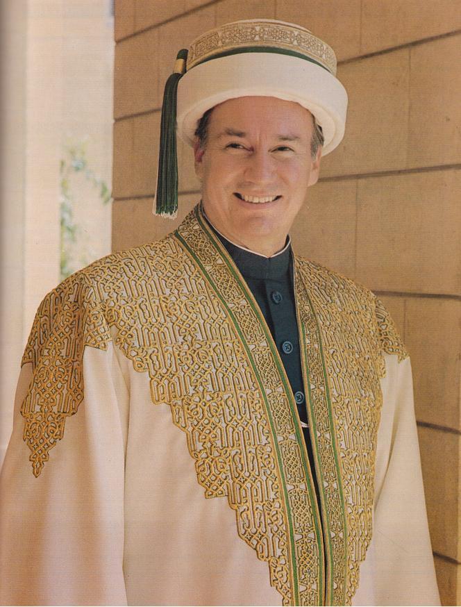 A smiling man dressed in an ornate traditional outfit featuring intricate gold embroidery, standing against a light-colored stone wall.