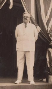 A historical black and white photograph of a man standing in formal attire, with arms relaxed at his sides, in front of draped fabric featuring a flag design.