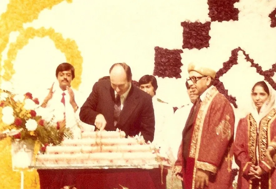 A historical birthday celebration featuring an Imam cutting a cake, surrounded by attendees in formal attire, with floral decorations in the background.