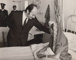 A man in a suit bending down to speak with a woman wearing a headscarf, seated on a hospital bed, with two men in the background.