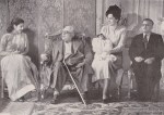 A historic black and white photograph of a group of five individuals seated in a room, with one person using a cane and holding a child, showcasing a family gathering.