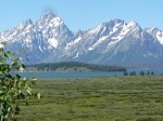 Snow-capped mountains in the background with a clear blue sky, surrounded by greenery and a calm body of water in the foreground.