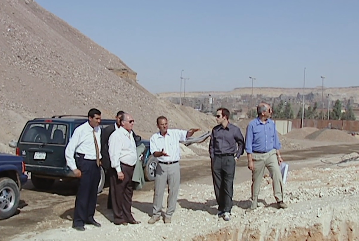 Five men in business attire stand on a construction site discussing plans, with vehicles parked nearby and a rocky landscape in the background.