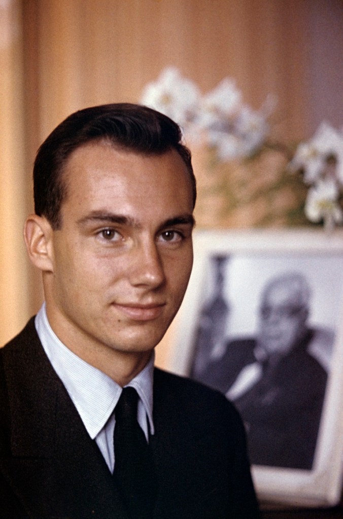 A young man in formal attire, smiling in the foreground, with a framed photograph of an older gentleman in the background, adorned with flowers.