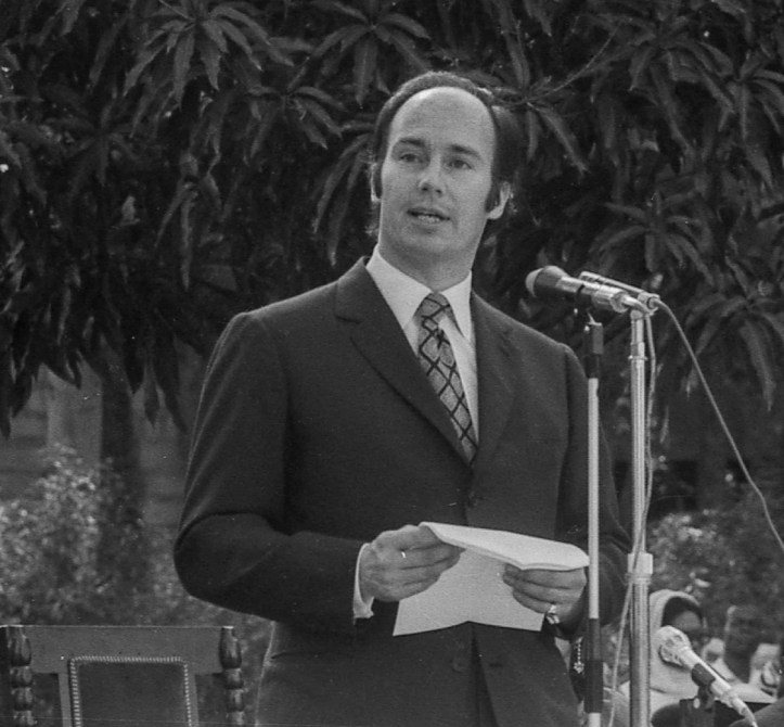 A man in a suit stands at a microphone outdoors, holding notes and speaking, with trees in the background.
