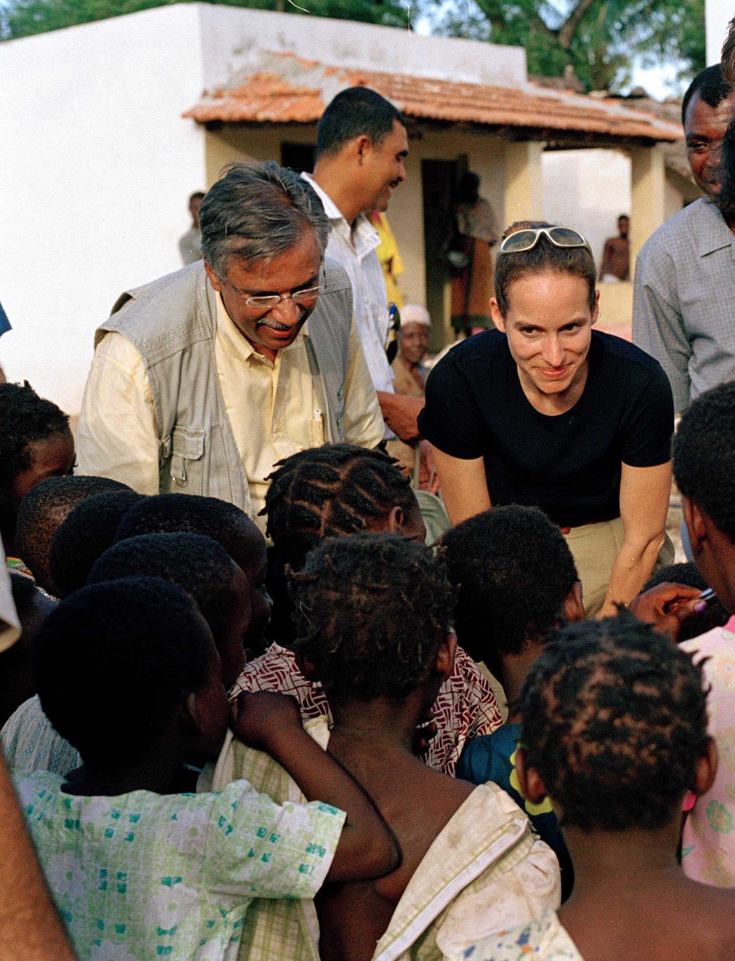 Princess Zahra Aga Khan interacts with children in a rural setting, accompanied by a man in a vest, amidst a group of onlookers.