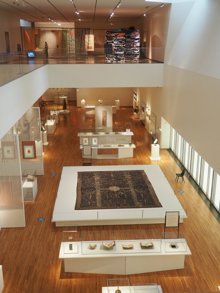 Interior view of a modern art museum showcasing various exhibits, including a prominently displayed rug, sculptures, and framed artworks. The space features large windows allowing natural light to illuminate the gallery.