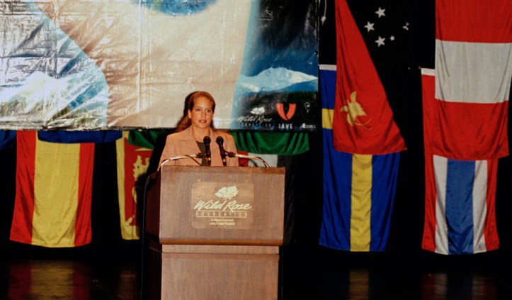 Princess Zahra Aga Khan speaking at an event with various international flags displayed in the background.