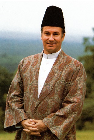 His Highness the Aga Khan, dressed in traditional attire, smiling and posing outdoors with a scenic backdrop.