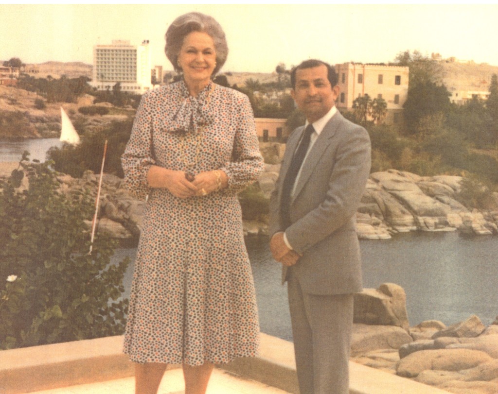 A woman in a floral dress standing beside a man in a suit, both smiling, with a river and buildings in the background.