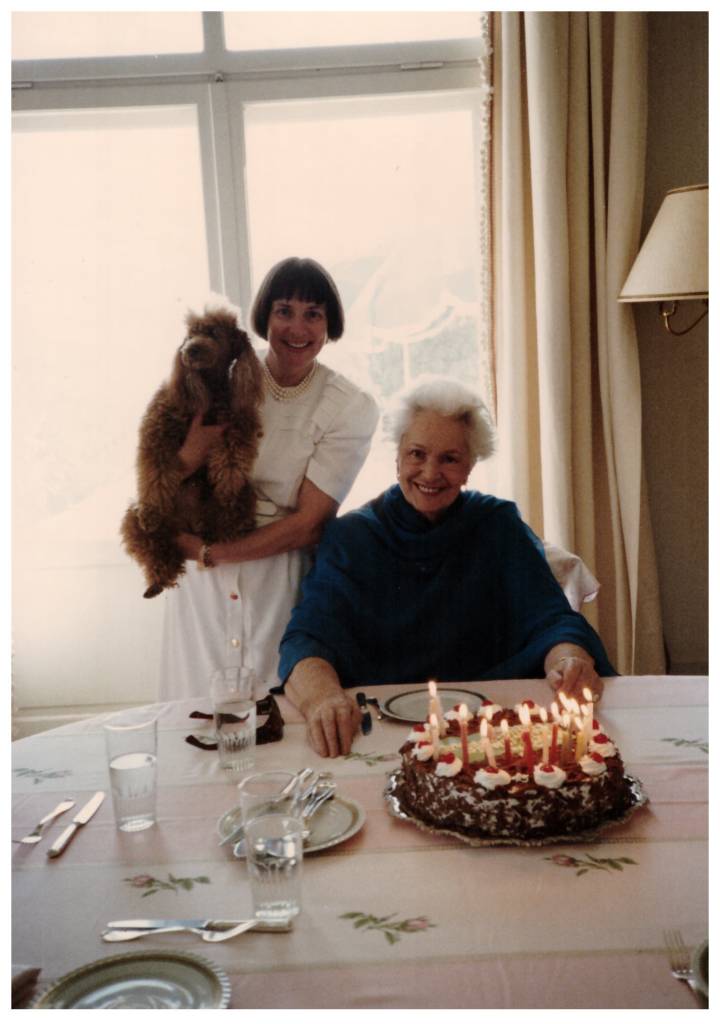 A woman holds a dog while standing next to another woman seated at a table with a birthday cake adorned with candles.