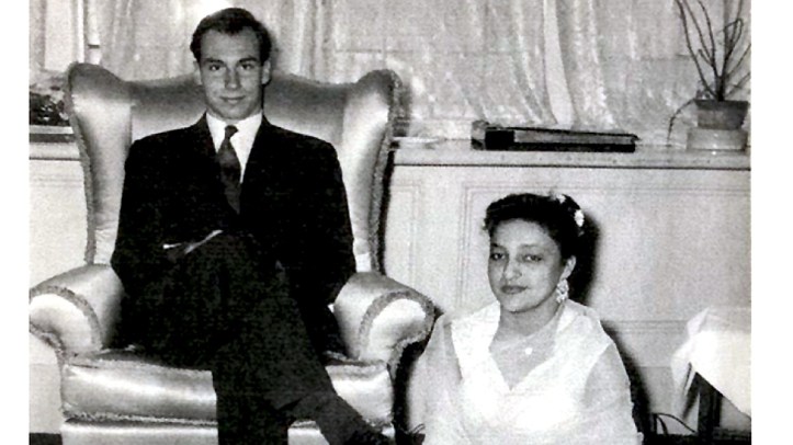 A black and white photograph featuring a man seated in a chair and a woman sitting on the floor beside him, both dressed elegantly. The setting appears to be an indoors with a softly lit background.