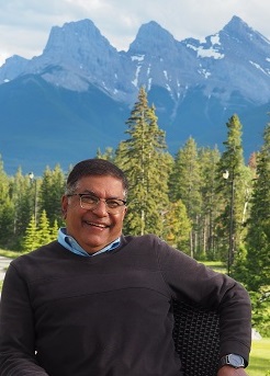 A smiling individual sitting on a chair surrounded by lush greenery and mountains in the background.
