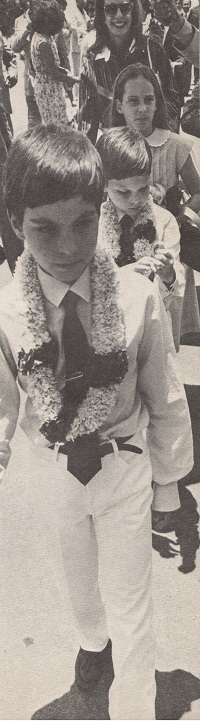 Black and white photograph of a group of children wearing flower garlands and formal attire. A boy is prominently featured in the foreground, with two other children visible behind him, while adults can be seen in the background.