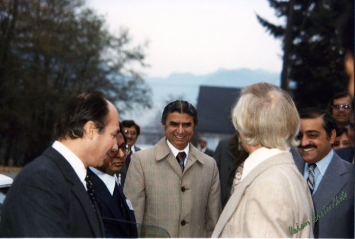 A group of men engaged in conversation outdoors, smiling and dressed in formal attire. The background features trees and a distant mountain range.