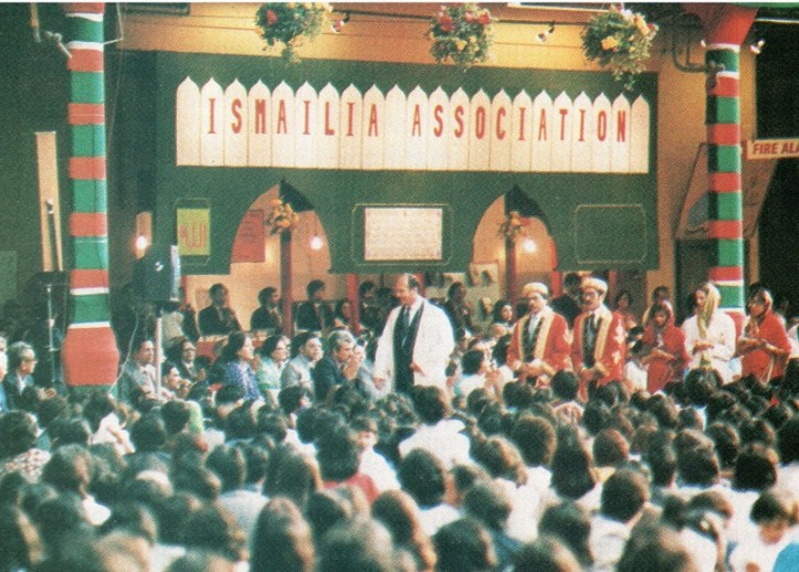 A gathering at the Ismailia Association showcasing a diverse audience seated on the floor, with speakers and performers on stage, under a decorated ceiling.