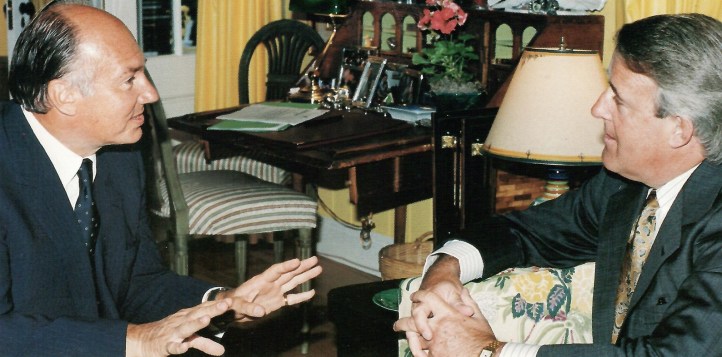 Two men engaged in conversation in a well-decorated room with a desk and flowers in the background.