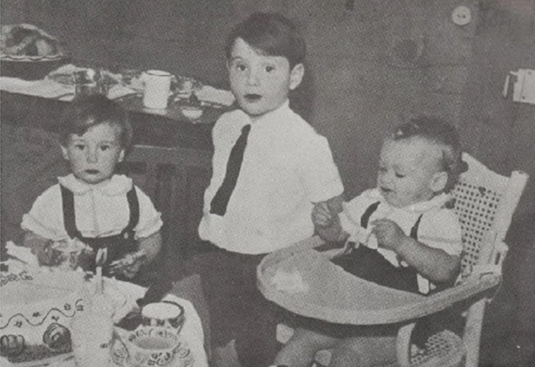 A historical black and white photograph of three young children sitting around a table. The oldest child, wearing a white shirt and a black tie, is positioned in the center, looking towards the camera. The younger children, both dressed in similar attire, are to his left and right, with one in a high chair.