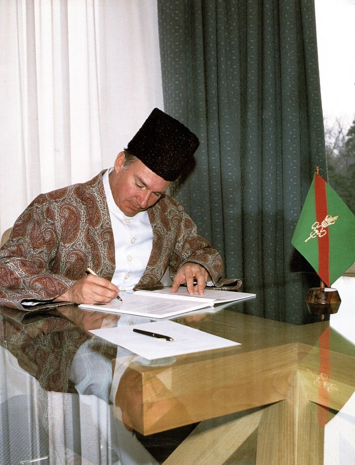 Mawlana Shah Karim, His Highness the Aga Khan IV, dressed in traditional attire, seated at a desk while signing documents, with a decorative flag beside him.
