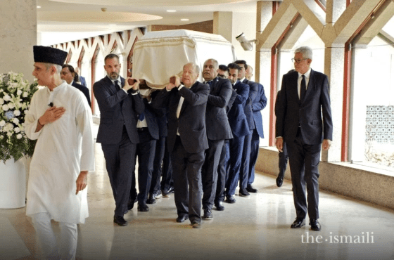 A solemn moment during a funeral procession, with individuals in formal attire carrying a coffin, while another person in traditional dress walks alongside.