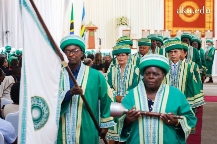 A ceremonial procession featuring individuals in traditional green and gold robes, with one person carrying a flag and another holding a staff, taking place during a graduation event.