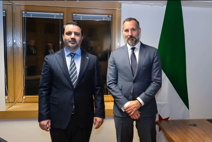 Two men stand together in a formal setting, facing the camera. One man is dressed in a dark suit with a striped tie, while the other is in a gray suit with a white shirt. A flag featuring red, white, and green can be seen in the background.