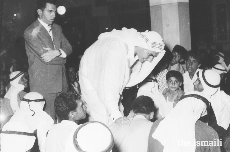 A black and white photograph of a crowd gathered around a man dressed in white traditional attire, who is bending down to speak with a seated group. The scene captures a moment of engagement and interaction, surrounded by onlookers, including children.