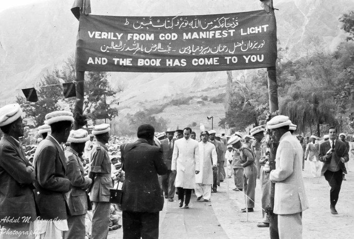A historic black and white photograph of a crowd gathered in a mountainous setting, with figures in traditional attire and military uniforms. A large banner is displayed above reading 'VERILY FROM COD MANIFEST LIGHT AND THE BOOK HAS COME TO YOU' in both English and Arabic. The focus is on a central figure walking towards the camera, while onlookers watch on.