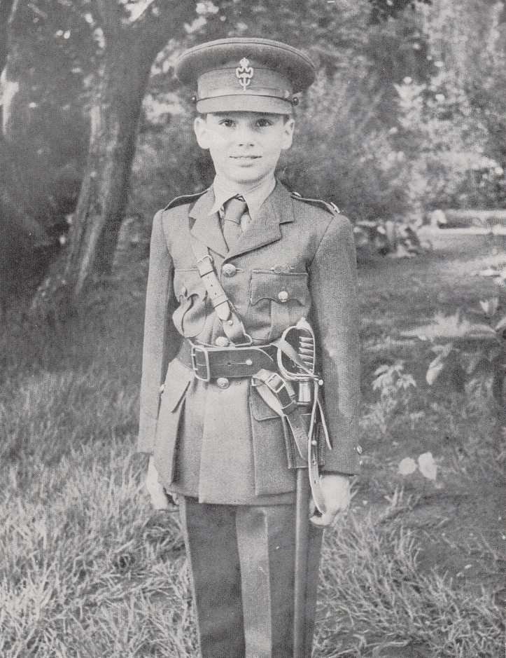 A young boy dressed in a military uniform, standing with a smile in a garden setting. He wears a hat and has a belt with a holster at his waist.