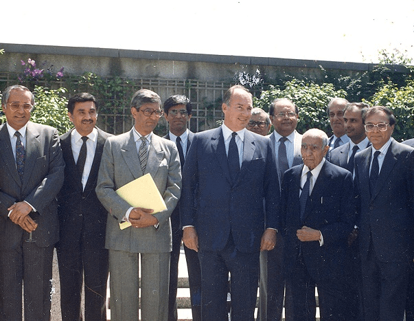Group photo of several men, including a prominent figure in a suit, standing outdoors in front of greenery.