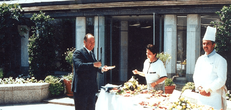 A man in a suit holds a piece of food while smiling at a woman in a black and white outfit, who is engaging with him at an outdoor dining setup. A chef in a white uniform stands nearby, with a table adorned with various dishes and flowers in the background.