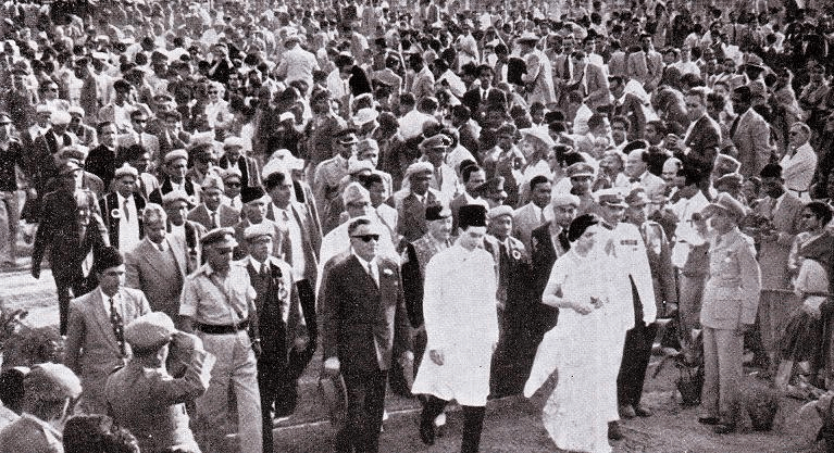 A historical black and white photograph featuring a large crowd gathered to witness an event, with prominent figures including a man in a formal suit and sunglasses walking alongside a woman in a white dress. Various individuals in military and formal attire are visible in the background, highlighting the significance of the gathering.