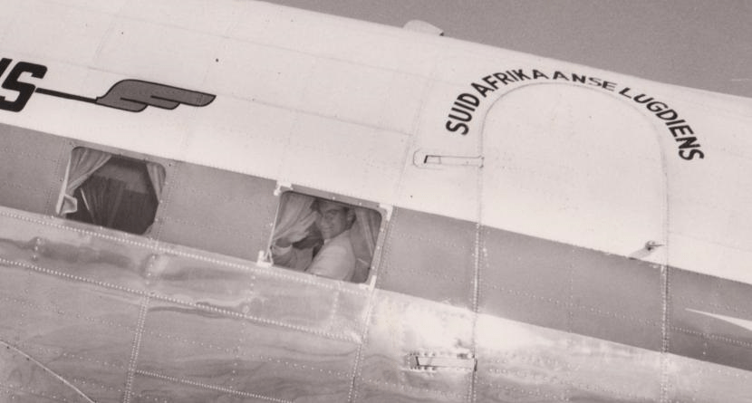 A historic black and white photograph of a man seated in the cockpit of an airplane, visible through a window decorated with the text 'SUID AFRIKAANSE LUGDIENS'.