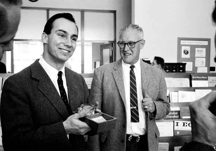 A black and white photograph of a man in a suit holding a small box with a smile, accompanied by another man in glasses and a suit, both appearing to discuss the contents.