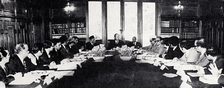A historical meeting taking place around a long table with several individuals engaged in discussion. The setting features a formal wooden interior with bookshelves in the background.