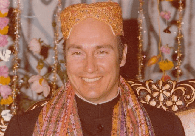 Mawlana Shah Karim, His Highness the Aga Khan IV, smiling while wearing a traditional cap and vibrant ceremonial attire against a backdrop of flowers and decorations.