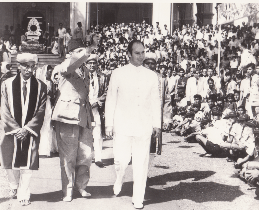 Mawlana Shah Karim walking among a crowd during a public event, surrounded by attendees in traditional attire and military uniforms.