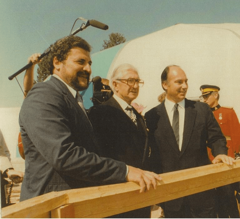 A group photo featuring three men at a ceremonial event, with two men smiling and standing on either side of a third man who appears to be receiving recognition, set against a backdrop of trees and a white structure.